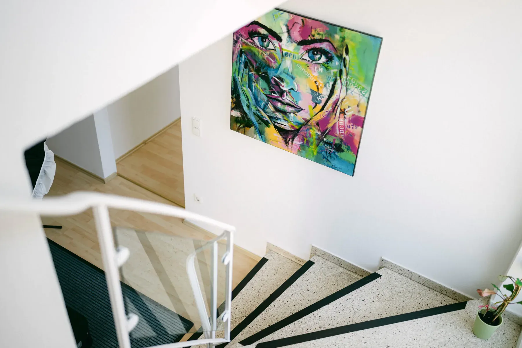 Ein Treppenabgang ist zu sehen. An der rechten Wand hängt ein buntes Gemälde, welches das Gesicht einer jungen Frau zeigt.