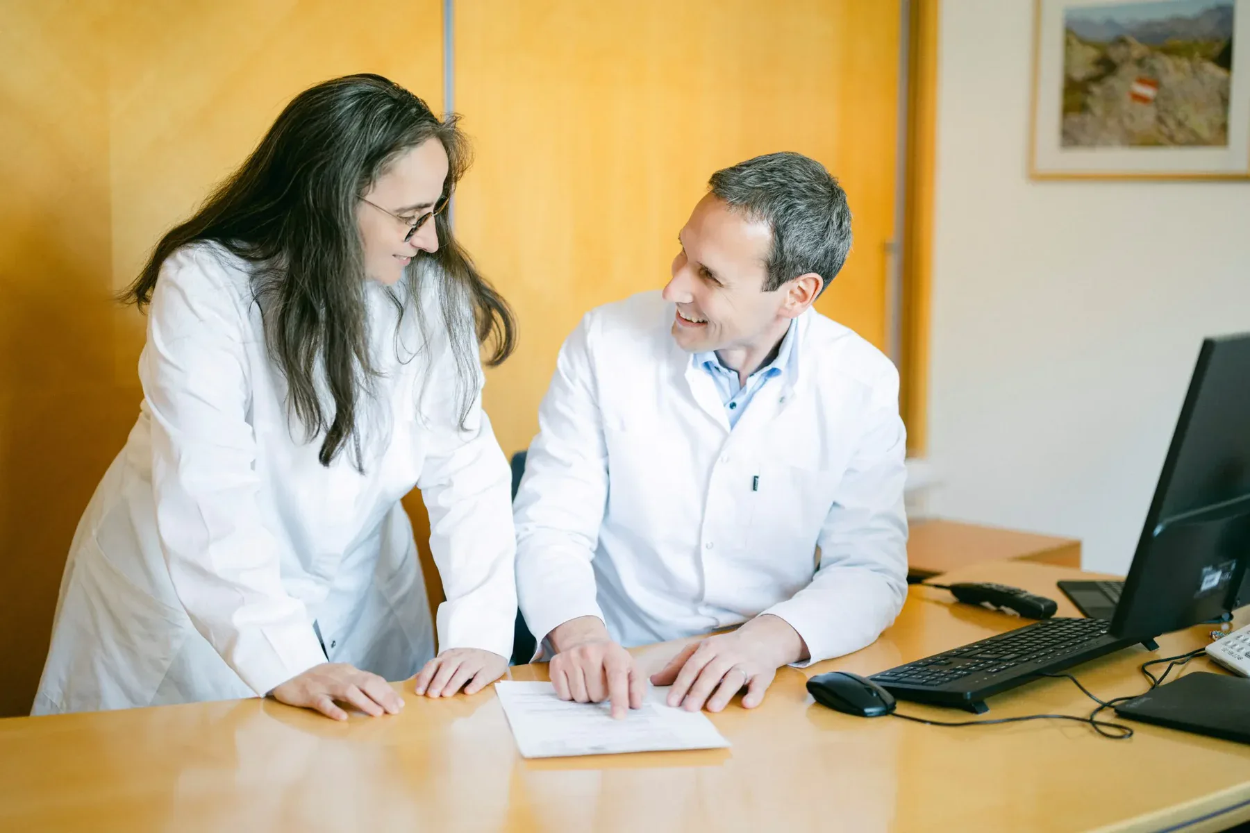 Beide Ärzte sitzen an einem Schreibtisch und diskutieren miteinander über einen Bericht, der ausgedruckt auf dem Tisch liegt.