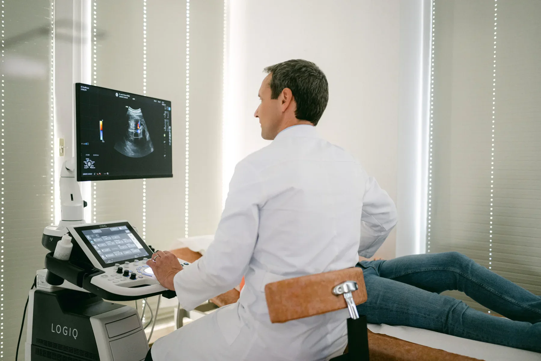 Dr. Olipitz führt einen Ultraschallscan bei einer Patientin durch, die neben ihm auf einer Liege Platz genommen hat.