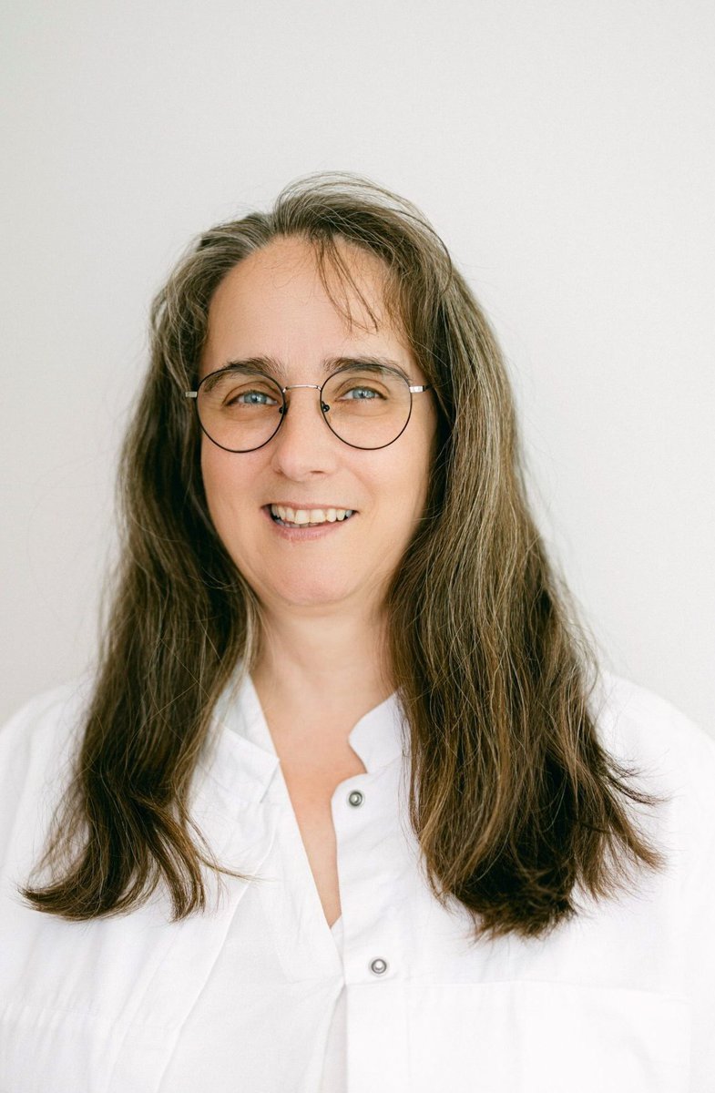 Frau Schäffer trägt einen weißen Arztkittel und blickt mit einem Lächeln in die Kamera. Sie trägt eine runde Brille und hat schulterlanges, braunes Haar.