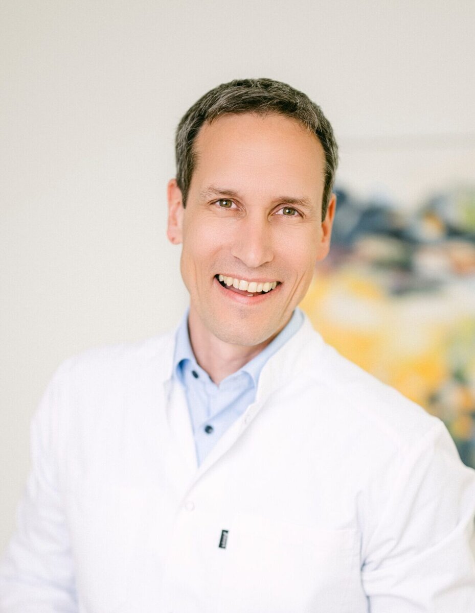 Herr Dr. med. Olipitz steht im Arztkittel vor einem Gemälde und lächelt in die Kamera.
