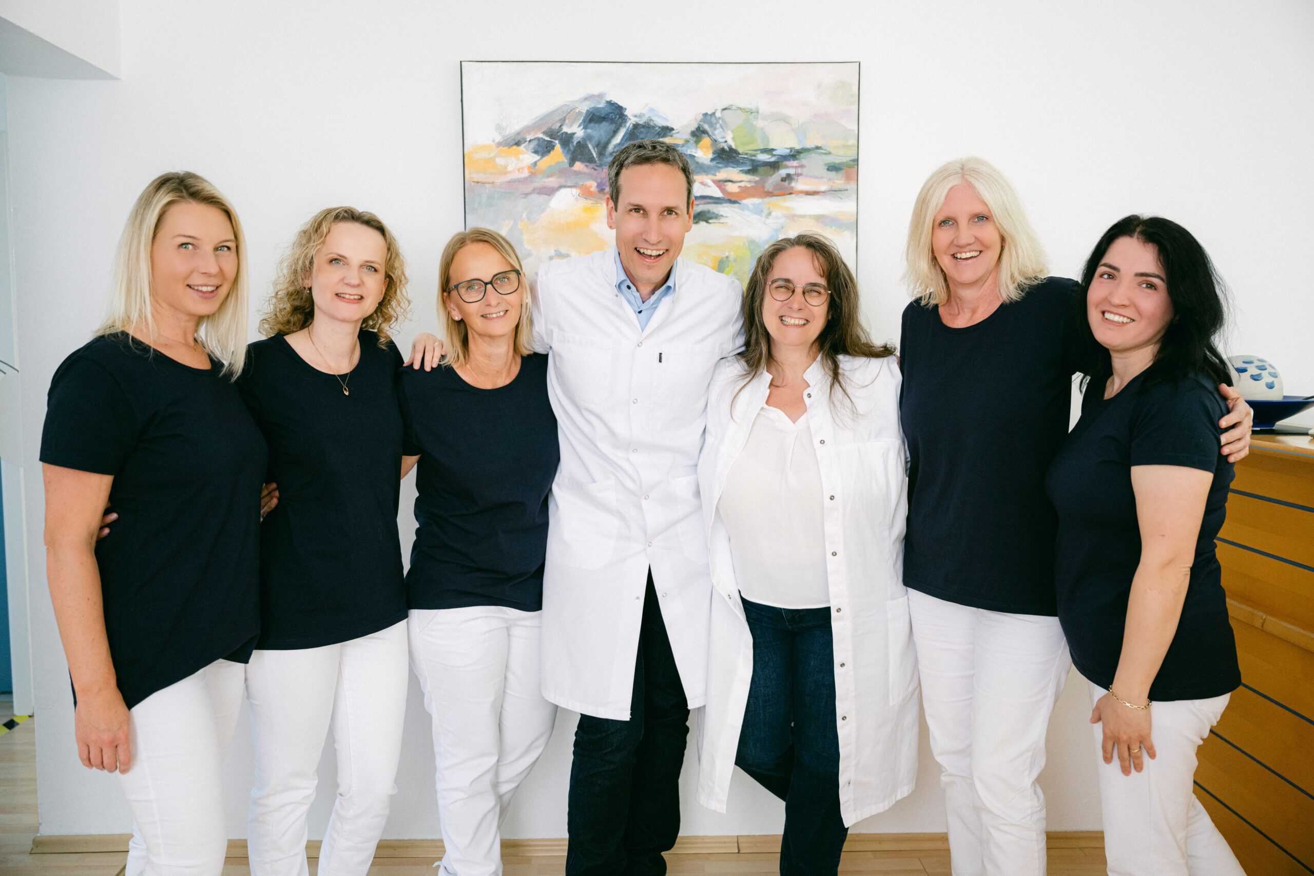 Das Team der Onkologie Altötting ist zu sehen. Die insgesamt sieben Personen stehen im Empfangszimmer der Praxis vor einem Gemälde und lächeln in die Kamera.
