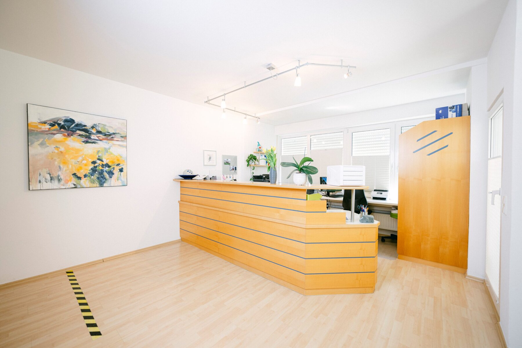 Der Empfangsraum der Praxis ist zu sehen. Ein großer Tisch mit mehreren Pflanzen befindet sich in der Mitte des Raumes und an der Wand hängen mehrere Bilder.